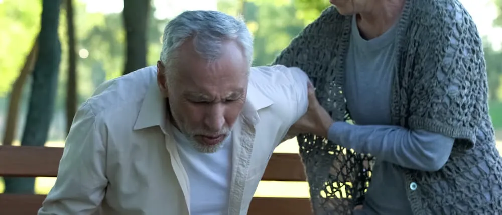 Woman comforting an older man in pain on a park bench after hernia mesh surgery